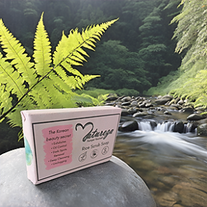 Narueza Rice scrub soap with pink packaging, is on the stone, near the lake and mountain and green leave is also behind the soap as well.